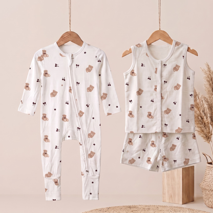 A baby clothing set displayed on wooden hangers against a soft beige background. The set includes a long-sleeve zip-front bamboo sleepsuit  and a matching sleeveless top and shorts set, both in white fabric with a repeating print of teddy bears and small black stars.