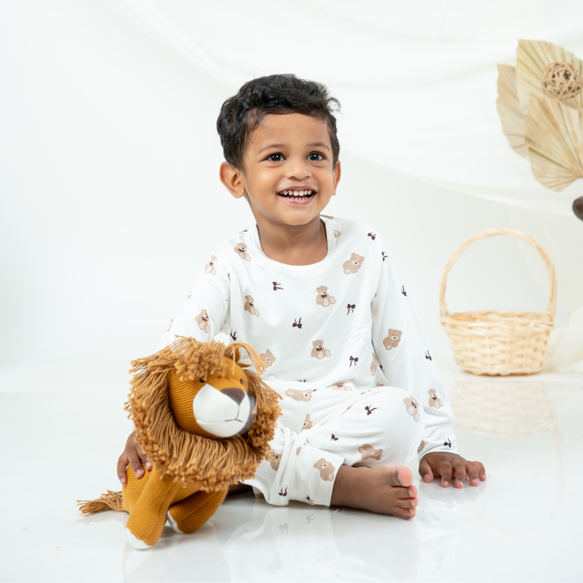 A smiling young child sitting on a white studio floor wearing a white long-sleeve bamboo lounge set with small teddy bear prints, holding a plush lion toy. A woven basket and dried palm leaves are softly blurred in the background.