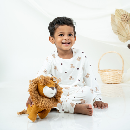 A smiling young child sitting on a white studio floor wearing a white long-sleeve bamboo lounge set with small teddy bear prints, holding a plush lion toy. A woven basket and dried palm leaves are softly blurred in the background.