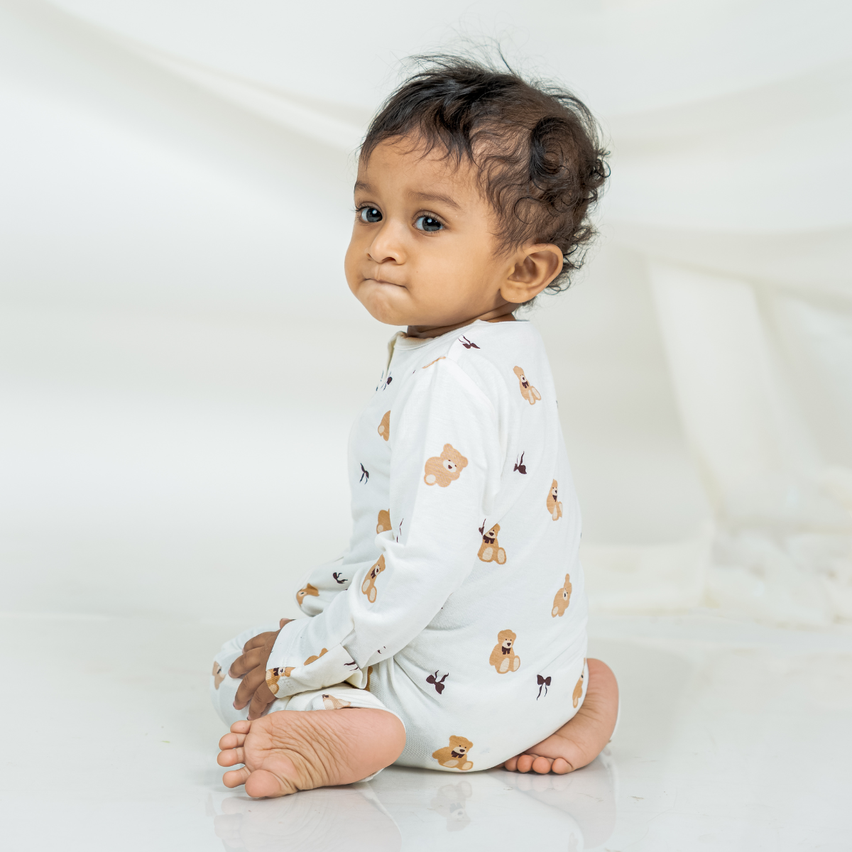 A baby sitting on a white studio floor wearing a white long-sleeve bamboo sleepsuit with a teddy bear print.