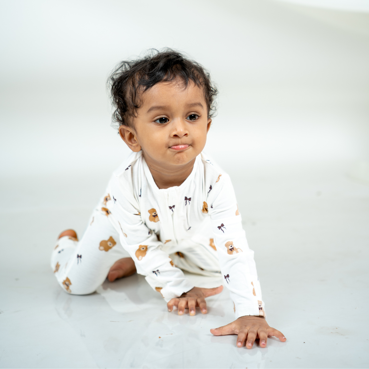 A baby crawling on a white studio floor wearing a bamboo sleepsuit with a small teddy bear pattern.