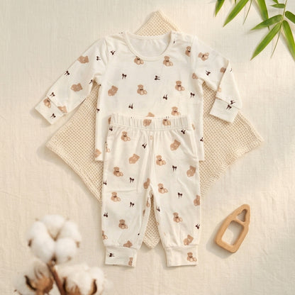 A baby two-piece bamboo lounge set consisting of a long-sleeve top and matching pants, laid flat on a textured cream blanket with decorative props including cotton stems, green leaves, and a wooden teething ring.