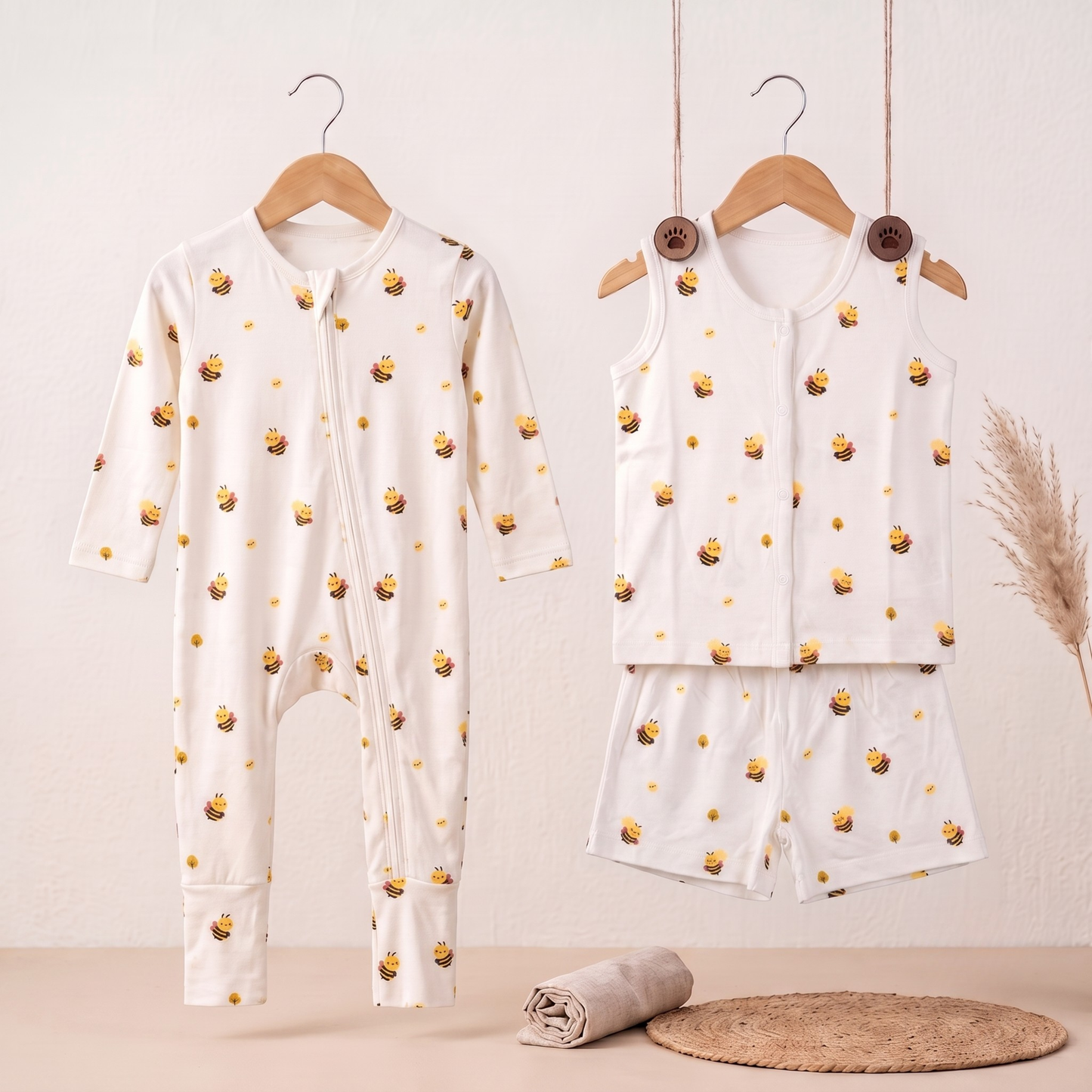A baby two-piece outfit set hanging on wooden hangers against a light beige wall. The set includes a long-sleeve zip-front bamboo sleepsuit and a matching sleeveless bamboo jabla and shorts set, both in white with a repeating yellow honeybee print.