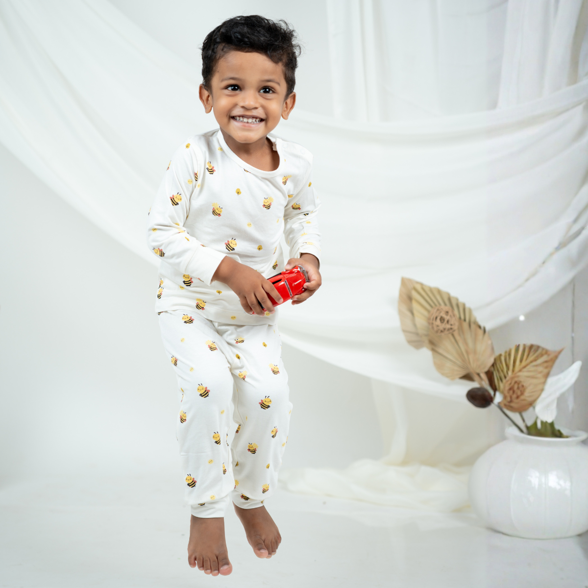A smiling young boy wearing a white long-sleevebamboo lounge set with small yellow and black bee prints, holding a small red toy against a soft white draped backdrop.
