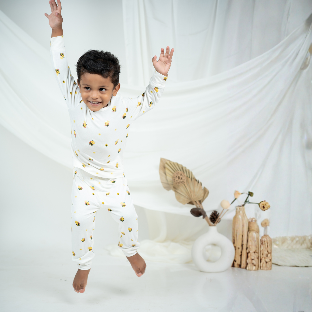 A young boy wearing a white long-sleeve bamboo lounge set with small yellow and black bee prints jumps in a playful pose against a white draped fabric backdrop.