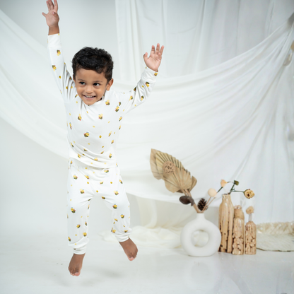 A young boy wearing a white long-sleeve bamboo lounge set with small yellow and black bee prints jumps in a playful pose against a white draped fabric backdrop.