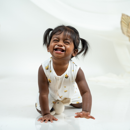 A young toddler girl with dark hair in pigtails is sitting on a white studio floor, smiling and laughing while wearing a bamboo jabla shorts set with a yellow and black bumblebee print.