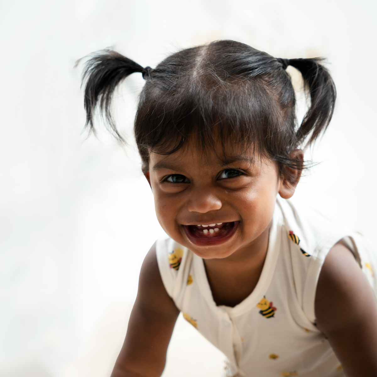 A close-up shot of a young child with dark hair in pigtails smiling, wearing a bamboo jabla shorts set with small yellow and black bee prints against a bright white background.