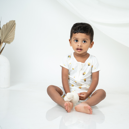 A toddler boy sitting cross-legged on a white studio floor, wearing a white short-sleeve bamboo romper with small yellow and black bee prints. He is holding a small white plush toy, with a white vase and dried leaf stems visible on the left side of the background.