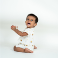 A baby sitting on a white studio floor wearing a white short-sleeve bamboo romper with small yellow and black bee prints, clapping their hands in a joyful expression.