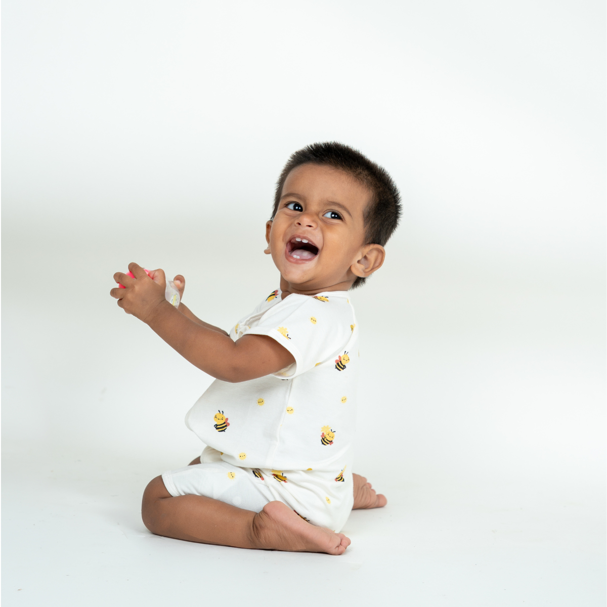 A baby sitting on a white studio floor wearing a white short-sleeve bamboo romper with small yellow and black bee prints, clapping their hands in a joyful expression.