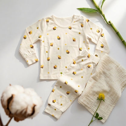 A baby two-piece bamboo lounge set laid flat on a white background, featuring a long-sleeve cream top and matching pants with a yellow bee print, alongside a soft cream muslin blanket. Decorative cotton boll, bamboo stem, and a small yellow flower are placed around the set.