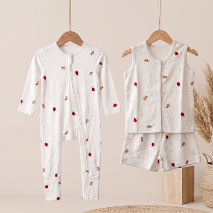 A baby bamboo sleepsuit and jabla shorts set displayed on wooden hangers against a warm beige background. The set includes a long-sleeve footless zip-front sleeper with a small red berry and light pink floral print, and a matching sleeveless vest and shorts set in the same fabric.