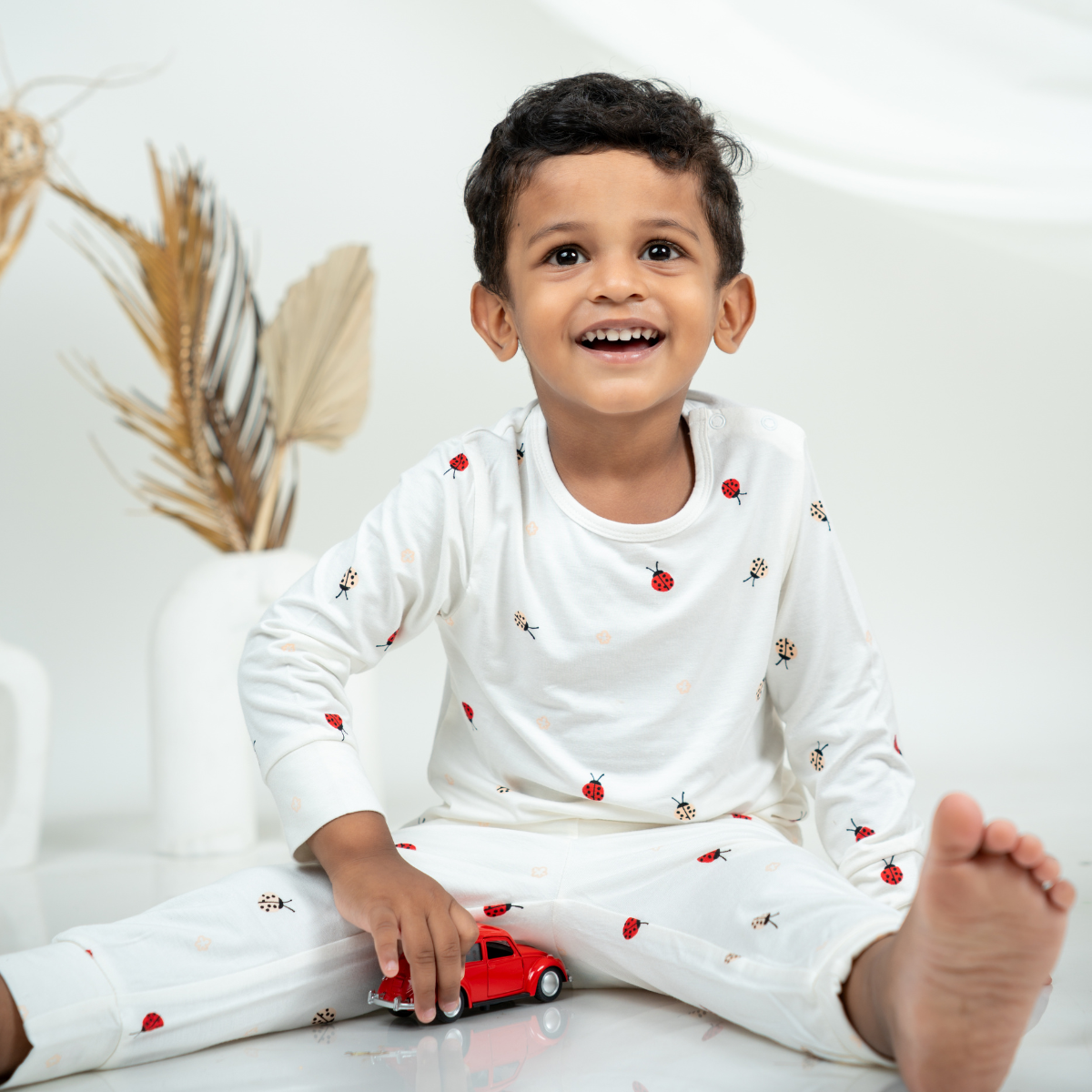 A young boy smiling while wearing a white long-sleeve bamboo lounge set with small red and black ladybug prints, sitting on a white floor and holding a small red toy car.