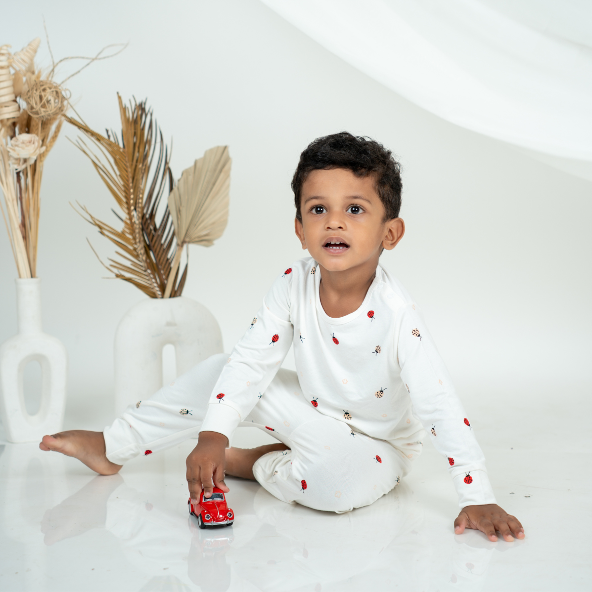 A young boy sitting on a white studio floor wearing a white bamboo lounge set with small red ladybug print. He is holding a small red toy car, with decorative dried plants in white vases in the background.
