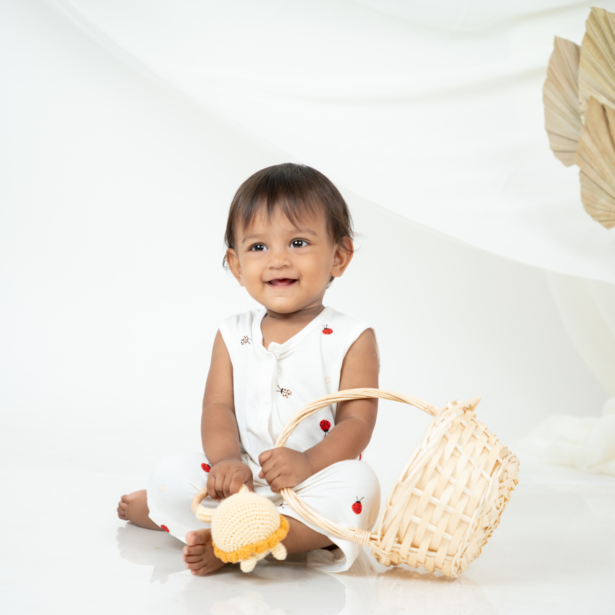 A baby sitting on a white studio floor wearing a white bamboo jabla shorts set with small red printed bug, holding a woven straw basket and a yellow-and-white plush toy.