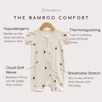 An off-white short-sleeve baby sbamboo romper with a front zipper and small red ladybug prints, hanging on a wooden hanger against a light beige background. The image features text callouts highlighting product features like 'Hypoallergenic', 'Thermoregulating', 'Cloud-Soft Weave', and 'Breathable Stretch'.