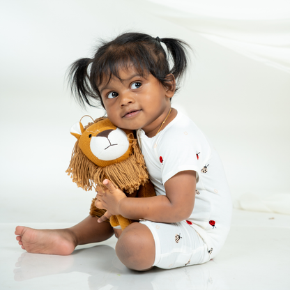 A toddler girl sitting on a white studio floor, wearing a white short-sleeve bamboo romper with small red and black printed ladybug motifs and holding a plush lion toy.