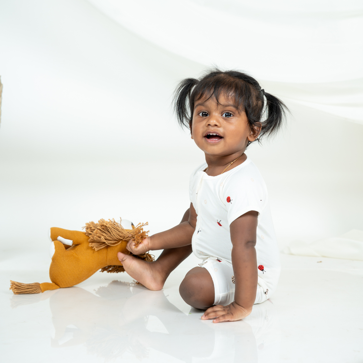 A toddler girl with dark hair in pigtails sits on a white studio floor, holding a mustard yellow plush toy. She is wearing a white short-sleeve bamboo romper with small red ladybug printed details.