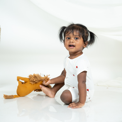 A toddler girl with dark hair in pigtails sits on a white studio floor, holding a mustard yellow plush toy. She is wearing a white short-sleeve bamboo romper with small red ladybug printed details.