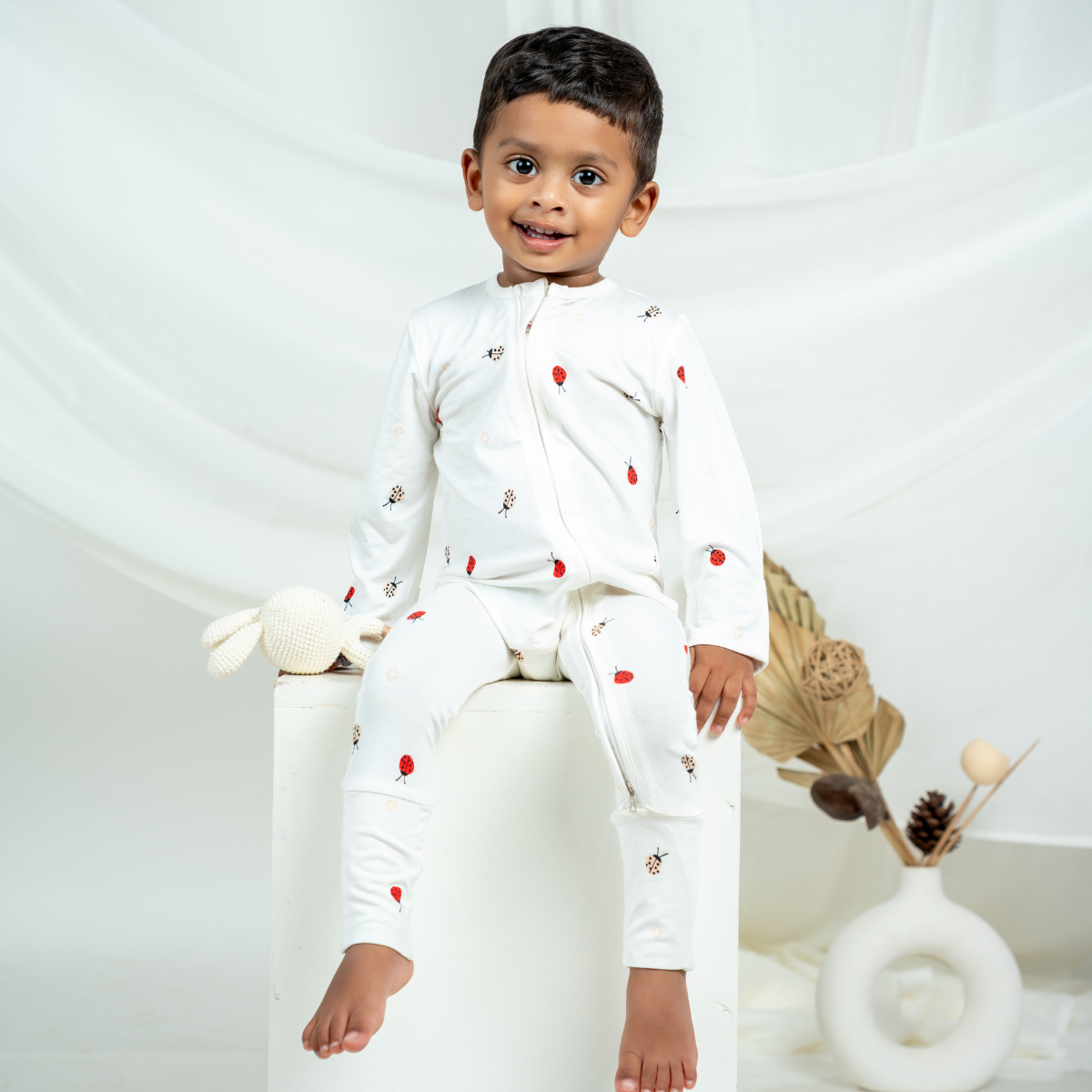 A young child sitting on a white pedestal wearing a white bamboo sleepsuit with small red ladybug print, photographed in a soft studio setting with draped white fabric and decorative dried flowers.