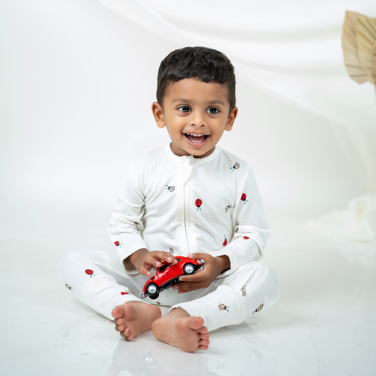A smiling young boy sitting cross-legged on a white studio floor, wearing a white bamboo sleepsuit with small red and black ladybug prints, while holding a red toy car.