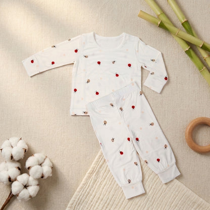 A baby two-piece bamboo lounge set laid flat on a textured beige fabric background, featuring a white long-sleeve top and matching pants with small red ladybug prints. Styled with cotton bolls, bamboo sticks, and a wooden ring toy.
