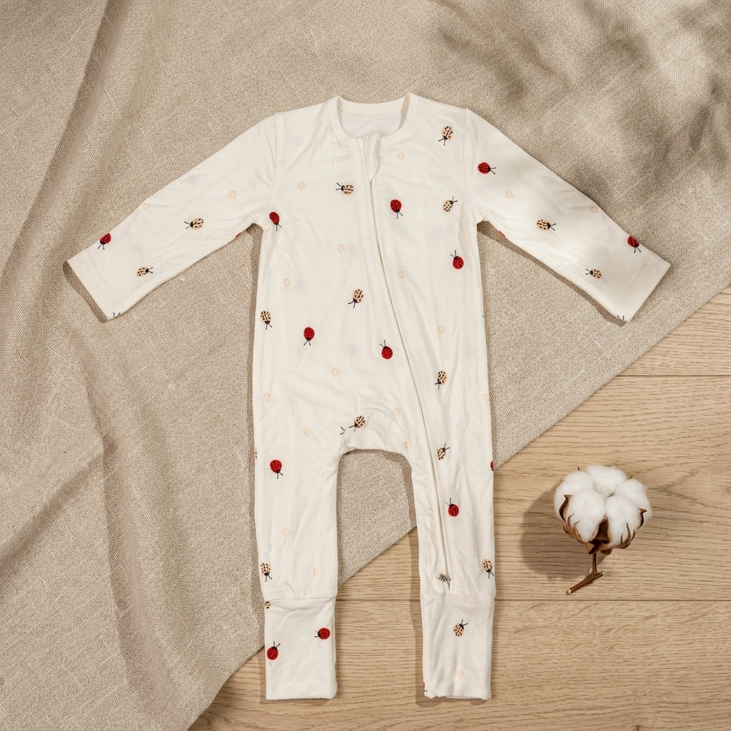 A baby long-sleeve bamboo sleepsuit in off-white, decorated with small printed red ladybugs, laid flat on a beige fabric backdrop over a light wood surface with a cotton boll prop.