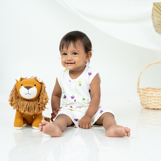 A baby sitting on a white studio floor next to a plush lion toy, wearing a white sleeveless bamboo jabla shorts set with small colorful fruit prints.