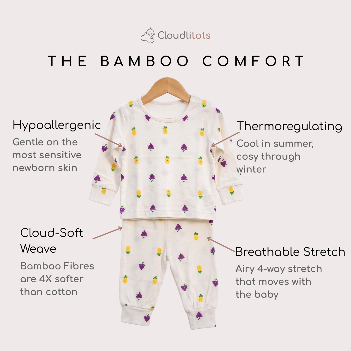 A baby bamboo lounge set displayed on a hanger against a light beige background, with text callouts describing its bamboo-comfort features. The set includes a long-sleeve top and matching pants in a warm off-white color, decorated with a small repeating pattern of grapes and yellow-green pineapples.
