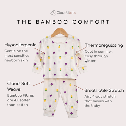 A baby bamboo lounge set displayed on a hanger against a light beige background, with text callouts describing its bamboo-comfort features. The set includes a long-sleeve top and matching pants in a warm off-white color, decorated with a small repeating pattern of grapes and yellow-green pineapples.