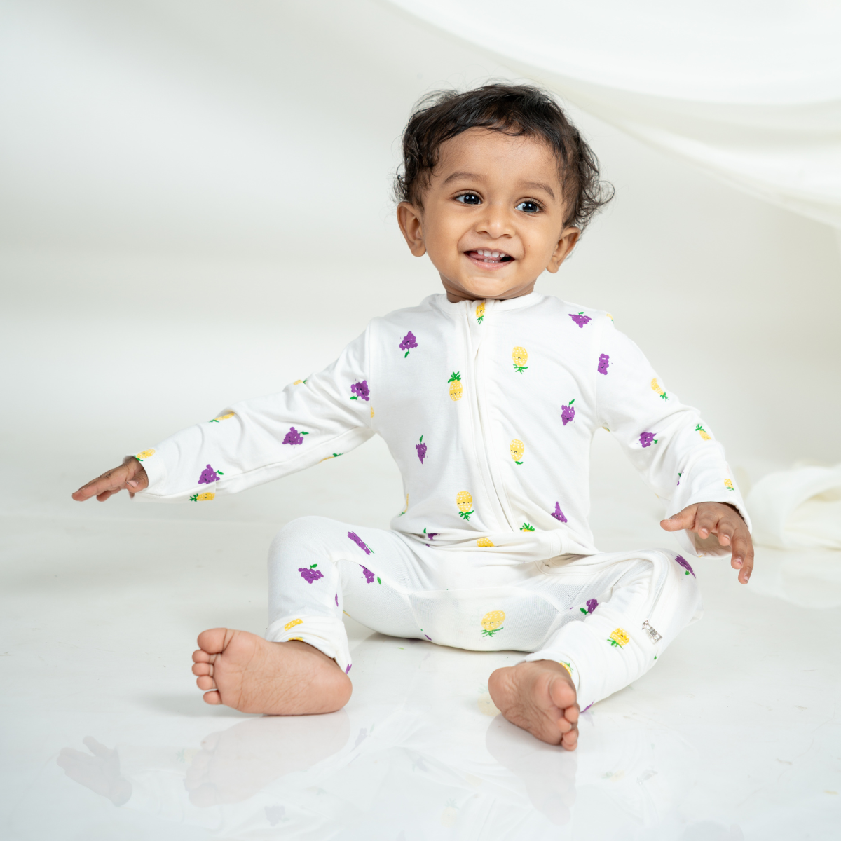 A smiling baby sitting on a white studio floor wearing a white bamboo sleepsuit with a small colorful fruits print.