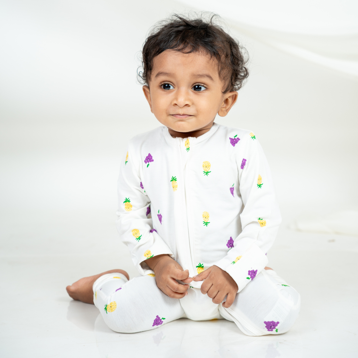A baby sitting cross-legged wearing a white bamboo sleepsuit with a small colorful pineapple and grape pattern, photographed against a clean white studio background.