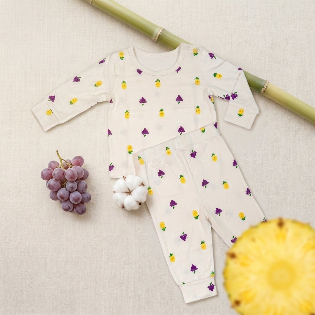 A baby long-sleeve bamboo lounge set in off-white fabric, decorated with a repeating pattern of small yellow pineapples and purple grapes. The set is styled in a flat-lay arrangement with a green bamboo stick, a bunch of purple grapes, a cotton boll, and a pineapple slice on a textured beige linen background.
