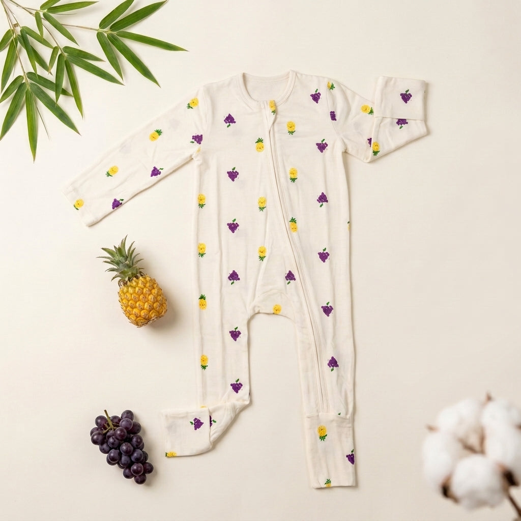 A baby bamboo sleepsuit laid flat on a light cream background, decorated with a yellow pineapple and purple grape pattern. Styled flat-lay props include a pineapple, a bunch of purple grapes, green leaves, and cotton bolls.