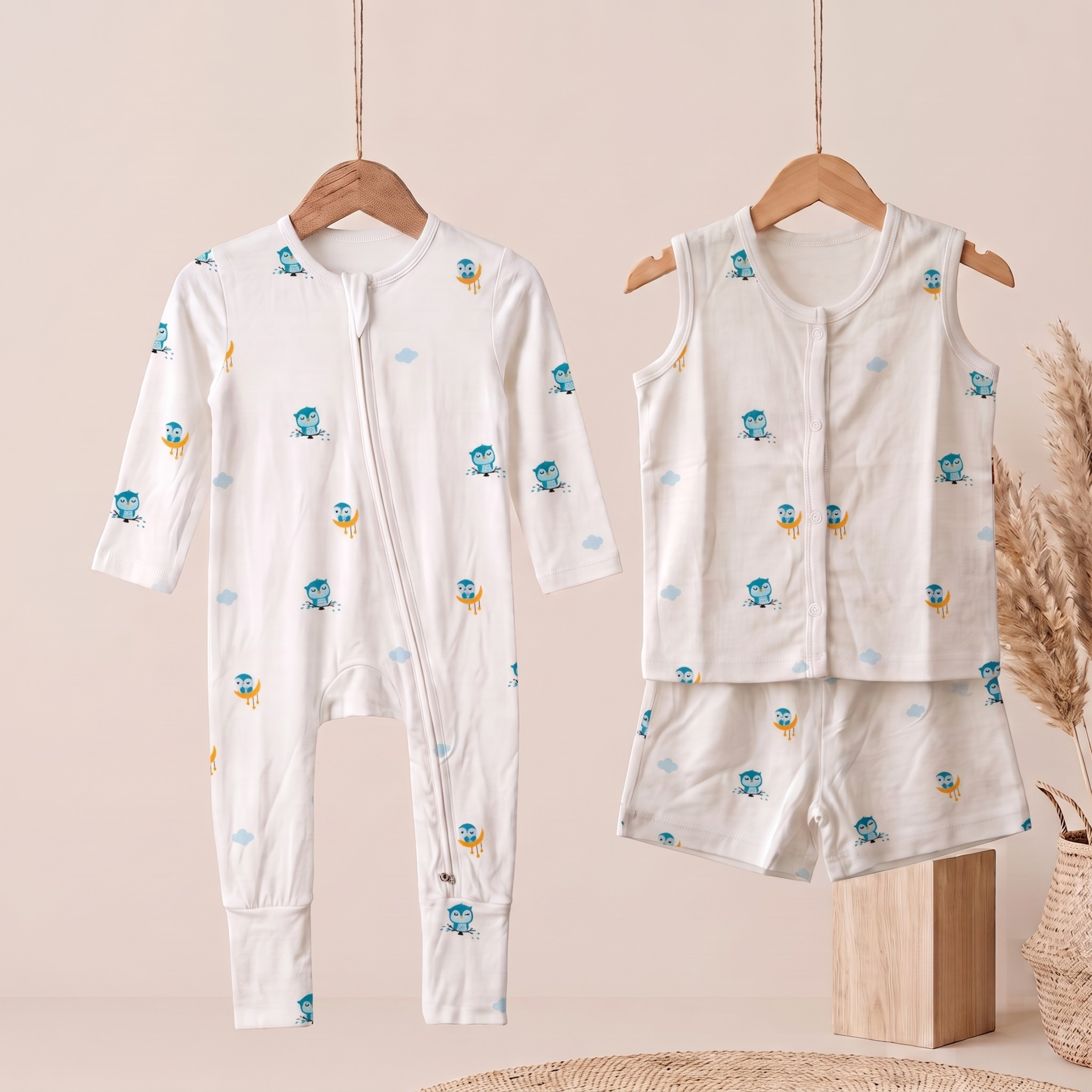 A baby bamboo sleepsuit and jabla shorts set displayed on wooden hangers against a warm beige background. The set includes a long-sleeve sleeper with a front zipper and a matching sleeveless top and shorts set, all in white fabric with a small blue and yellow owl cartoon cloud print.
