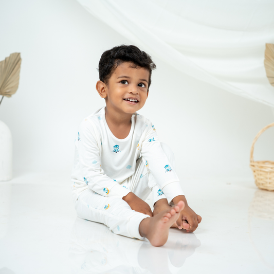 A smiling young boy sits cross-legged on a glossy white floor, wearing a white long-sleeve bamboo lounge set with small blue and yellow owl cartoon prints. The background is a bright white studio setting with soft draped fabric and minimal decor.