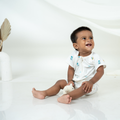 A smiling baby sitting on a glossy white floor, wearing a white short-sleeve bamboo romper with small blue and yellow owl prints. The baby is holding a small white stuffed animal.