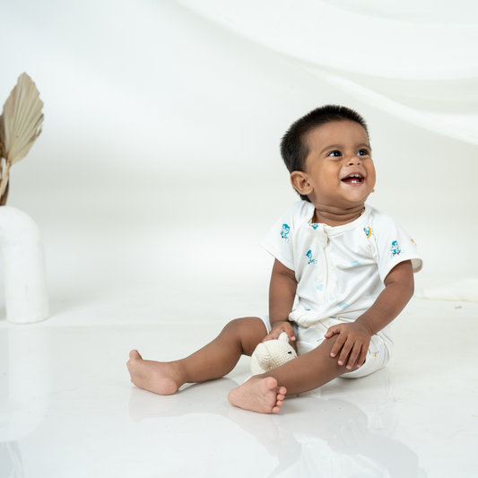 A smiling baby sitting on a glossy white floor, wearing a white short-sleeve bamboo romper with small blue and yellow owl prints. The baby is holding a small white stuffed animal.