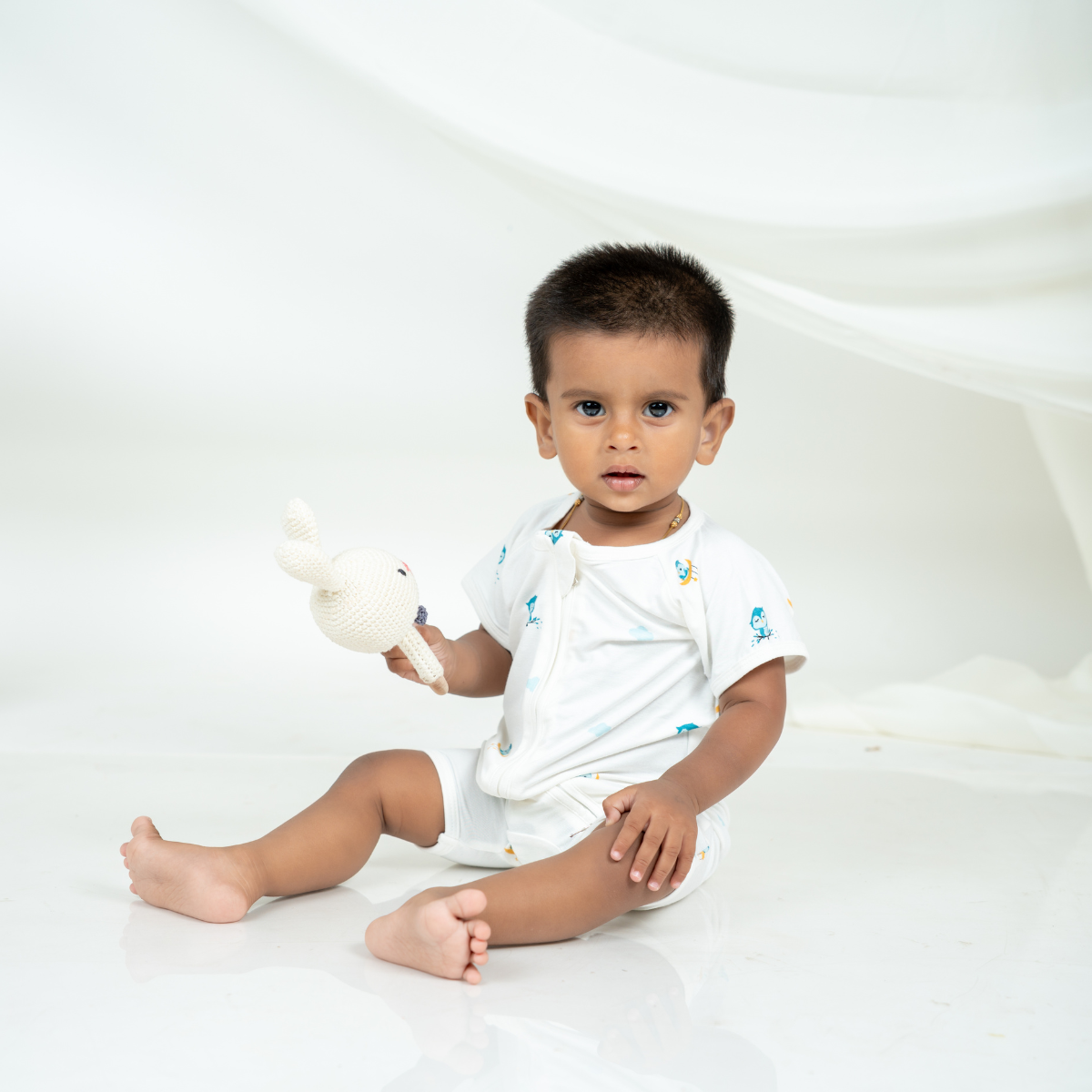 A baby sitting on a white studio floor holding a white plush toy, wearing a white short-sleeve bamboo romper with small colorful owl illustrations.