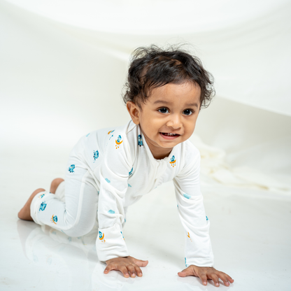 A smiling toddler crawling on a white surface wearing a white bamboo sleepsuit with small blue and yellow owl prints.