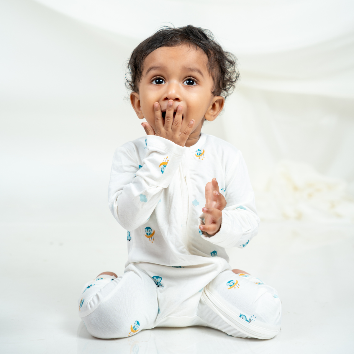 A baby sitting cross-legged wearing a white bamboo sleepsuit with small colorful cartoon prints, photographed against a soft white studio background.