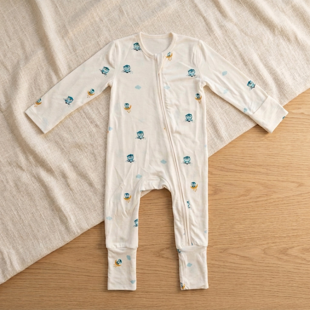 A baby long-sleeve bamboo sleepsuit  in off-white, decorated with small blue cartoon owl characters and light blue clouds, laid flat on a light wood surface with a beige blanket.