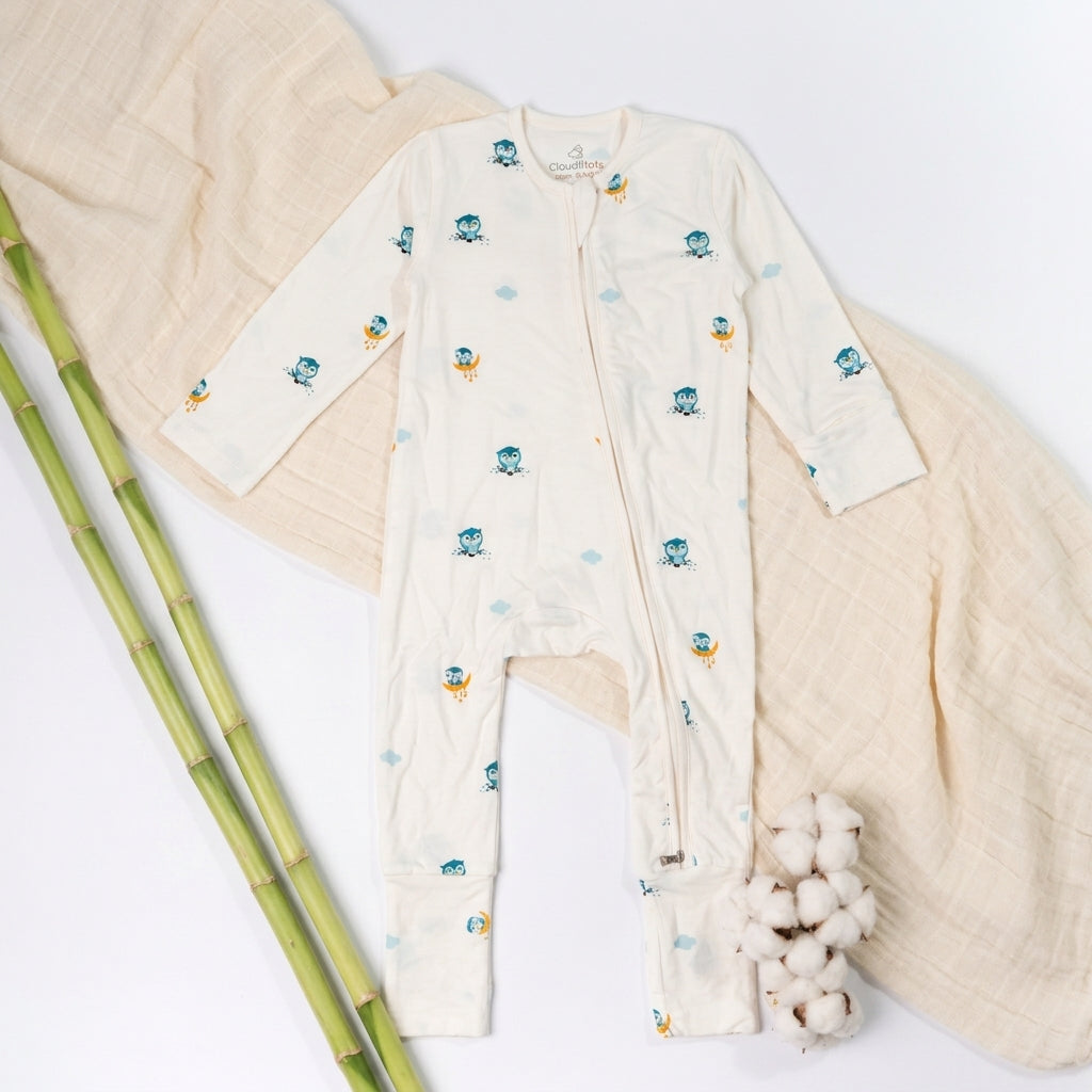 A baby long-sleeve bamboo sleepsuit  in off-white, decorated with small blue cartoon owl characters and light blue clouds, laid flat on a light beige blanket.