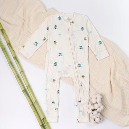 A baby long-sleeve bamboo sleepsuit  in off-white, decorated with small blue cartoon owl characters and light blue clouds, laid flat on a light beige blanket.
