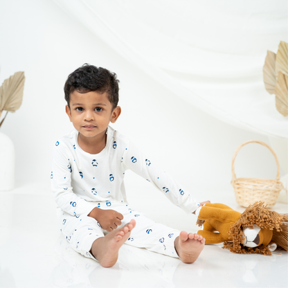 A young child sitting on a white floor wearing a white long-sleeve bamboo lounge set with small blue printed penguin motifs. The child is posed next to a plush lion toy and a woven basket, with a soft white draped backdrop behind them.