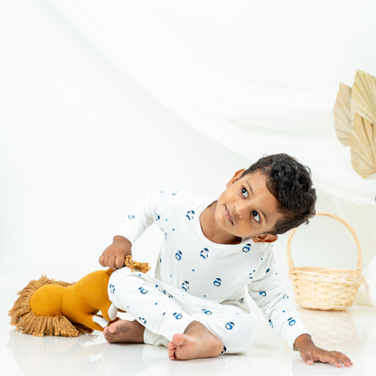 A young boy wearing a white long-sleeve bamboo lounge set with small blue penguin prints sits on a white floor, holding a yellow plush lion toy. A woven basket is visible in the background against a bright white studio setting.