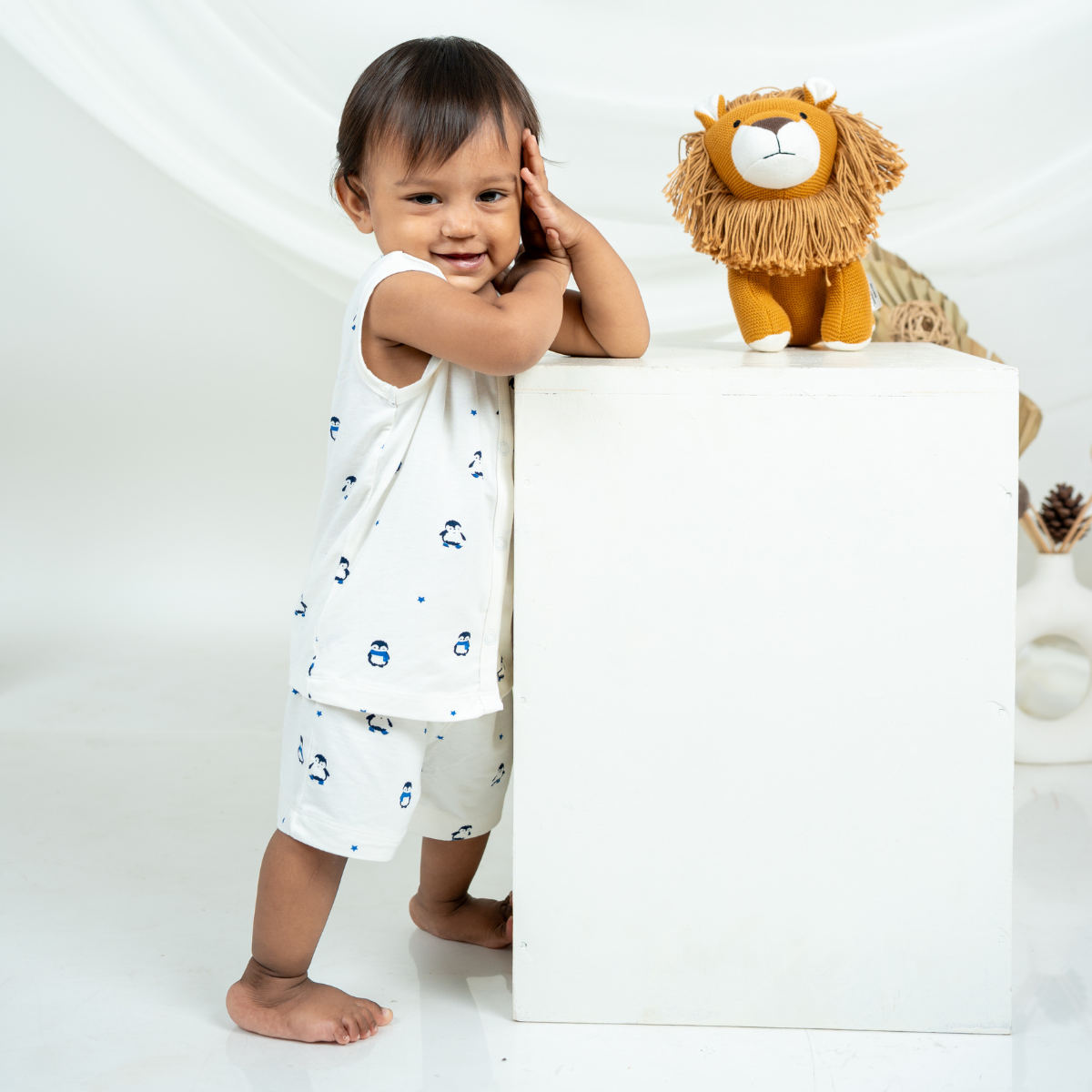A toddler stands barefoot beside a white pedestal in a bright studio setting, wearing a bamboo jabla shorts set with small blue cartoon penguin motifs. A plush lion toy sits on top of the pedestal.