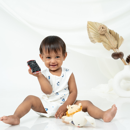 A toddler sitting on a white studio floor wearing a bamboo jabla shorts set with small blue penguin prints, holding a small black toy car. A crocheted stuffed animal lies on the floor nearby, with decorative dried palm leaves in a white vase in the background.