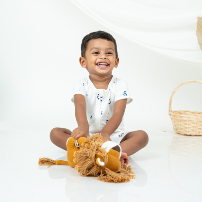 A smiling toddler boy sits cross-legged on a white studio floor, holding a plush lion toy. He is wearing a white short-sleeve bamboo romper with small blue penguin cartoon motifs.